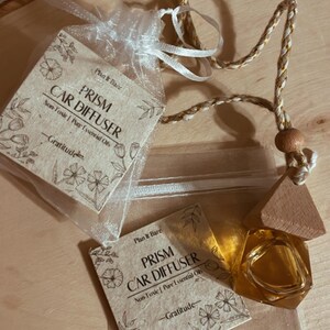 May include: A set of two car diffusers with a wooden charm. The diffusers are labeled "Prism Car Diffuser" and are made with pure essential oils. The charm is a triangular piece of wood with a gold-colored glass bottle attached.