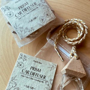 May include: A wooden car diffuser with a clear glass bottle and a gold string. The diffuser is labeled "Prism Car Diffuser" with the text "Non-Toxic | Pure Essential Oils" and "Lift Off" on a white background.