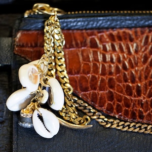 May include: A gold chain with a charm made of white seashells hangs from a brown leather purse with a black zipper.