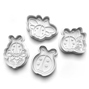 May include: Four white plastic cookie cutters in the shape of ladybugs and related designs. One cutter is a ladybug on a flower, another is a ladybug with wings, and two others are ladybugs with different details.