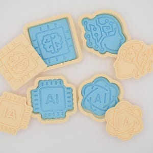 Puede incluir: Colección de cortadores de galletas beige claro y azul en varias formas, incluyendo un cerebro, una placa de circuito y las letras "AI". Los cortadores de galletas están dispuestos sobre una superficie blanca, sugiriendo un tema de tecnología o inteligencia artificial.