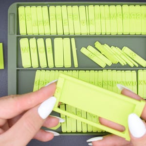 Könnte beinhalten: Ein hellgrünes Alphabet-Stempelset mit einzelnen Buchstabenstempeln und einer Ablage zur Aufbewahrung. Die Stempel sind in Reihen angeordnet, mit einem Rahmen in der Hand. Der Hintergrund ist eine dunkelgraue Oberfläche.