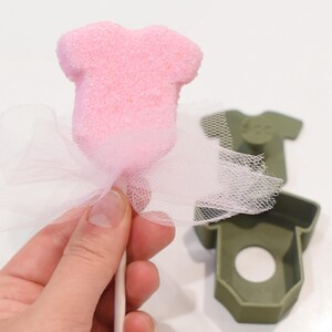May include: A pink, sugar-coated, baby onesie-shaped treat on a stick, adorned with a white tulle bow. A green plastic onesie-shaped mold is in the background. The treat is held by a person.