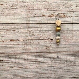 Puede incluir: Un pincho decorativo de cuentas y metal con un lazo en la parte superior. El pincho presenta un cubo de madera, cuentas doradas, moteadas y con dibujos blancos. El pincho está sobre una superficie de vetas de madera clara.