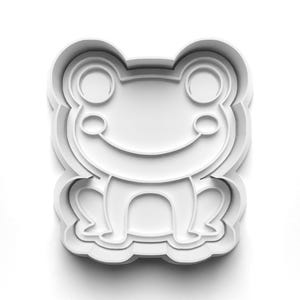 May include: A white plastic frog-shaped cookie cutter. The cutter features a smiling frog with large eyes and detailed legs. The design is made of layered plastic, creating a three-dimensional effect. Ideal for baking themed cookies.