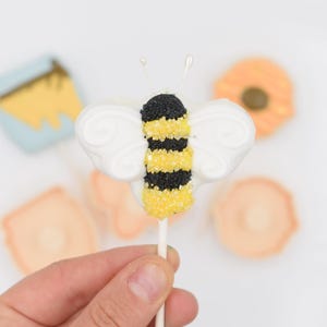 以下が含まれることがあります： 手で持った、棒の上の蜂の形をしたケーキポップ。 蜂は白い羽と黒と黄色のスプリンクルで飾られた体を持っています。 他のぼやけたお菓子が背景にあります。