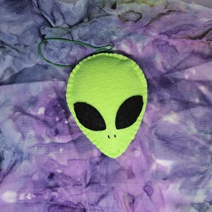 May include: Green felt alien ornament with black eyes. The ornament is hanging from a green string.