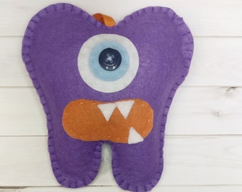 May include: A purple felt tooth-shaped monster with a large eye, orange mouth, and white teeth. The eye has a blue and white iris with a black button center. An orange ribbon is attached at the top.
