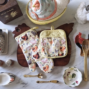 Könnte beinhalten: Ein Set aus zwei gelben und weißen Ofenhandschuhen mit einem Hühnermuster. Die Handschuhe liegen auf einem Holzbrett mit einem Hahnendesign. Die Handschuhe sind perfekt, um Ihre Hände beim Kochen oder Backen zu schützen.