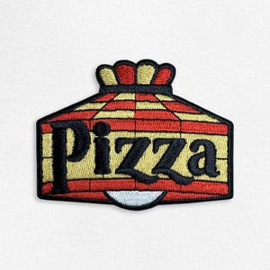 May include: Embroidered patch featuring the word "Pizza" in bold black letters. The patch has a red and yellow brick pattern design with a crown-like top. A white semi-circle is at the bottom of the patch.
