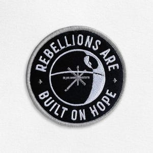May include: A black and white embroidered patch with the text "Rebellions are built on hope" and a stylised image of the Death Star.