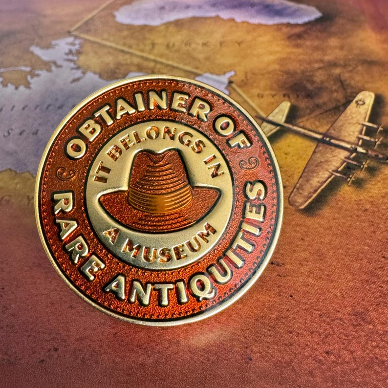 Pode incluir: Uma moeda redonda, cor de bronze, com a inscri&ccedil;&atilde;o "OBTAINER OF RARE ANTIQUITIES" e "IT BELONGS IN A MUSEUM". A moeda apresenta um chap&eacute;u fedora no centro. O fundo &eacute; um mapa com um modelo de avi&atilde;o.