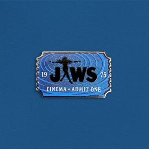 Könnte beinhalten: Eine rechteckige Emaille-Anstecknadel in Form einer alten Kinokarte mit gewelltem Rand. Das Design zeigt einen blauen Hintergrund mit konzentrischen Wellenmustern, den schwarzen Schriftzug JAWS, die Zahlen 19 und 75 sowie CINEMA ADMIT ONE.
