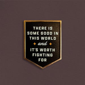 Può includere: Spilla in smalto nero e oro con la citazione "THERE IS SOME GOOD IN THIS WORLD and IT'S WORTH FIGHTING FOR" in lettere bianche. La spilla ha un bordo dorato ed è posta su uno sfondo marrone scuro.