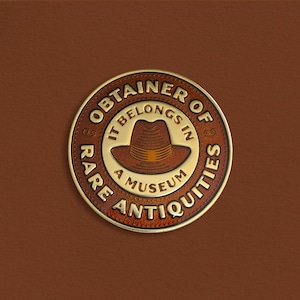 Pode incluir: Um emblema circular marrom e dourado com o texto "OBTAINER OF RARE ANTIQUITIES" e "IT BELONGS IN A MUSEUM" em torno de uma ilustra&ccedil;&atilde;o de fedora marrom. O emblema est&aacute; sobre um fundo marrom semelhante ao couro.