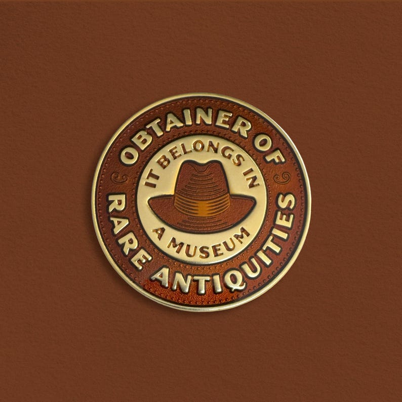 Pode incluir: Um emblema circular marrom e dourado com o texto "OBTAINER OF RARE ANTIQUITIES" e "IT BELONGS IN A MUSEUM" em torno de uma ilustra&ccedil;&atilde;o de fedora marrom. O emblema est&aacute; sobre um fundo marrom semelhante ao couro.
