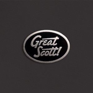 May include: A black and silver oval pin with the text "Great Scott!" in a stylized font.