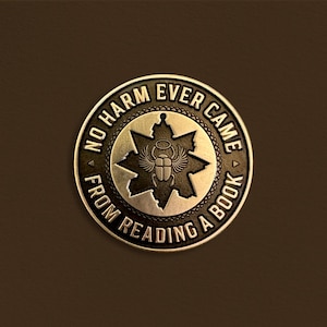 May include: A circular bronze-toned metal coin or medallion featuring a central embossed scarab beetle with wings and a halo, set against a star-shaped background. The text NO HARM EVER CAME FROM READING A BOOK is engraved along the outer border.