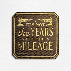 May include: A square embroidered patch with rounded corners featuring a dark brown background and gold thread embroidery. The text reads IT'S NOT the YEARS IT'S THE MILEAGE, flanked by two small gold winged emblems at the top and a horizontal line below.