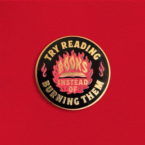 Pode incluir: Um pin redondo de esmalte preto e dourado com o texto "TRY READING BOOKS INSTEAD OF BURNING THEM". A palavra "BOOKS" está em letras douradas, rodeada por chamas. O pin está em um fundo vermelho.