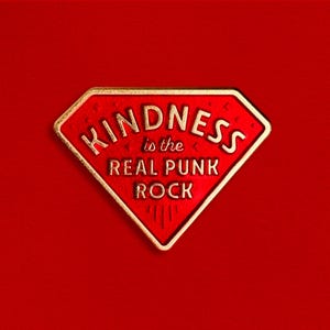 May include: A red diamond-shaped badge with gold edging, featuring the gold text "KINDNESS is the REAL PUNK ROCK". The badge is set against a plain red backdrop.
