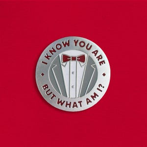 Pode incluir: Pin prateado com uma gravata borboleta vermelha e um gráfico de smoking. O texto ao redor do gráfico diz "Eu sei que você é, mas quem sou eu?"