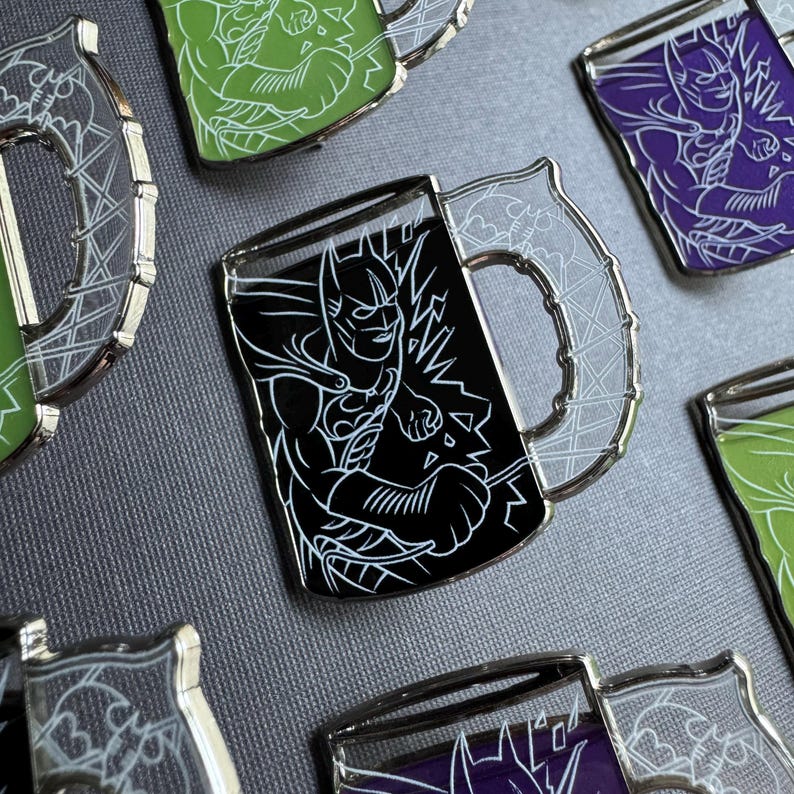 May include: Enamel pins in the shape of mugs, featuring superhero designs. The pins have a black background with white line art of a superhero. The mug handles and rims are silver. Other pins have green and purple backgrounds.