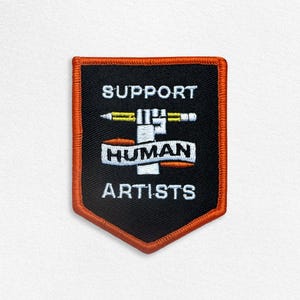May include: A shield-shaped embroidered patch with a black background and an orange border. It features a white graphic of a hand holding a yellow pencil, with a white banner reading HUMAN across it, and the text SUPPORT and ARTISTS in white.
