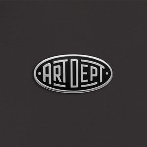 May include: Oval-shaped silver metal emblem with the words "ART DEPT" in a bold, stylized font. The emblem is set against a dark gray, textured background, creating a striking contrast. The design is clean and modern.