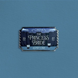 Op de afbeelding: Een metalen speld in de vorm van een bioscoopkaartje. De speld toont de tekst "The Princess Bride" en "Admit One". De achtergrond is een blauwe gradiënt met een zilveren rand. De speld heeft ook de tekst "Cinema" en de nummers "19" en "87".