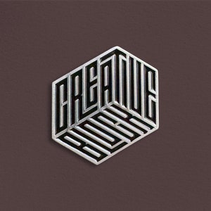 May include: A silver and black metal pin with the word "CREATIVE" spelled out in a 3D, geometric design.