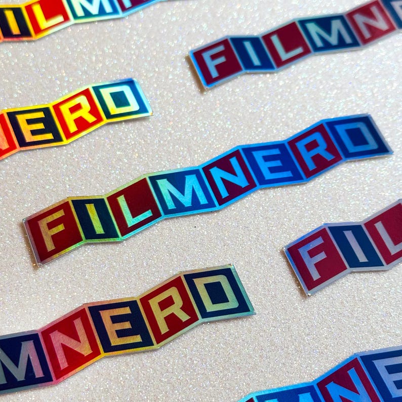 May include: A set of five iridescent stickers with the words "FILM NERD" spelled out in individual blocks. The stickers are holographic and have a rainbow effect.