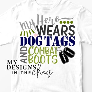 Puede incluir: Camiseta blanca con un diseño azul marino y verde que dice "My Hero... Wears Dog Tags and Combat Boots" con dos chapas de perro que dicen "Daddy" y "Mommy". El diseño también incluye el texto "My Designs in the Chaos".