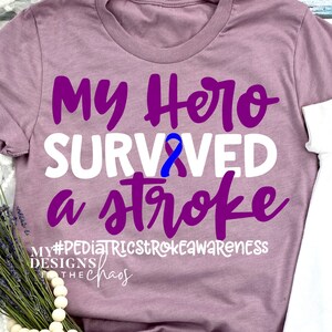 Peut inclure: T-shirt mauve avec l'inscription "My Hero Survived a Stroke" en violet et blanc. Un ruban bleu est au centre. Le texte "#PediatricStrokeAwareness" et "Designs by the Chaos" sont en bas.