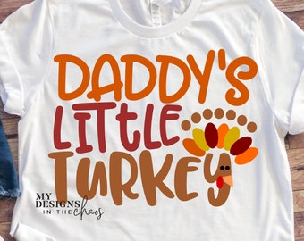 Puede incluir: Camiseta blanca con el texto "Daddy's Little Turkey" en letras naranja, rojo y marrón. Un pavo de dibujos animados con una papada roja y plumas naranja, amarillo y rojo está a la derecha del texto. El texto "My Designs in the Chaos" está en la esquina inferior izquierda.