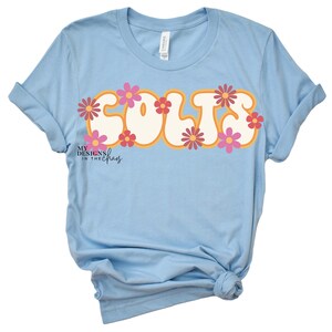 May include: Light blue t-shirt with the word "COLTS" in a retro font, outlined in orange and decorated with pink and orange flowers. The shirt has rolled-up sleeves and a knotted hem. The brand "MY DESIGNS IN THE CHAOS" is printed at the bottom.