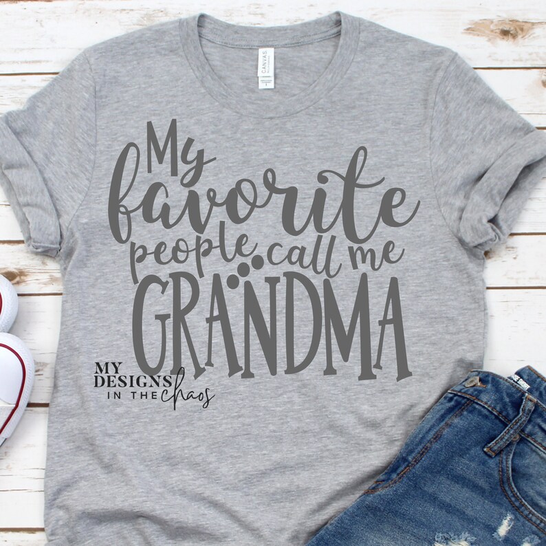 以下が含まれることがあります： グレーのTシャツに、グレーの筆記体で「My favorite people call me GRANDMA」という文字が書かれています。メインのテキストの下には、より小さなフォントで「MY DESIGNS IN THE CHAOS」という文字が印刷されています。