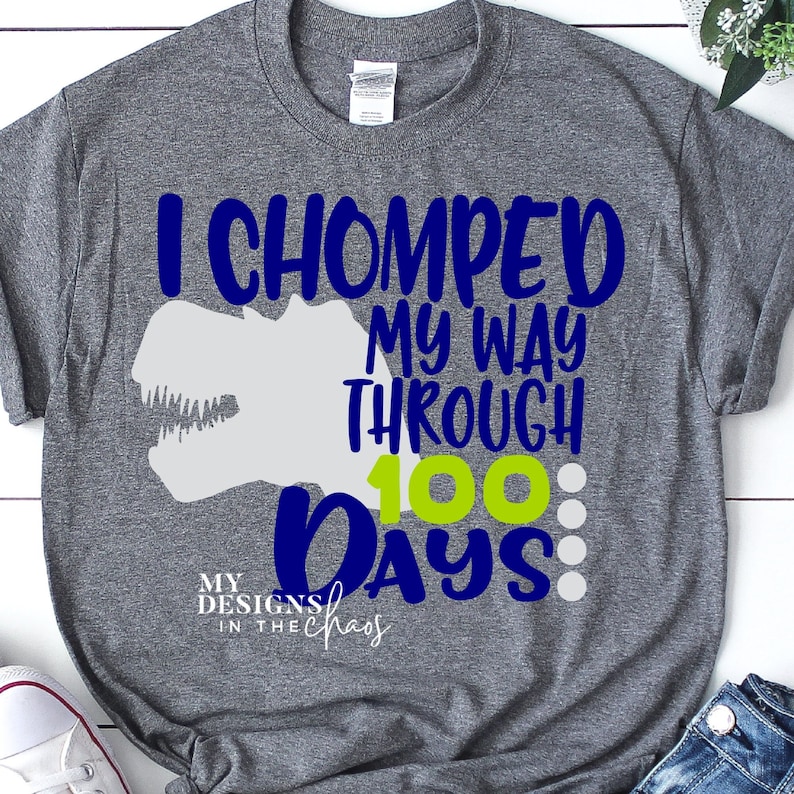 Pode incluir: Camiseta cinza com um gr&aacute;fico de dinossauro azul e o texto "Eu comi meu caminho atrav&eacute;s de 100 dias". O texto est&aacute; em azul e verde. A camiseta tem o texto "My Designs in the Chaos" no canto inferior esquerdo.