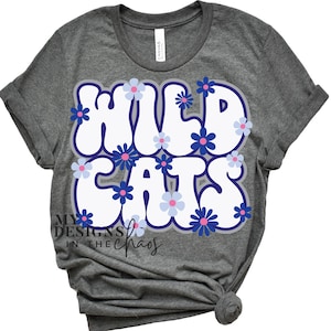 May include: A dark gray t-shirt with the words "WILD CATS" in white bubble letters outlined in purple. The letters are decorated with blue and pink flower graphics. The shirt has rolled-up sleeves and a knot at the bottom.