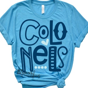 May include: A light blue t-shirt with the word "Colonels" in navy blue, with a white heart and dots. The shirt is made of a soft material and has rolled-up sleeves. The brand "Canvas" is visible on the tag.