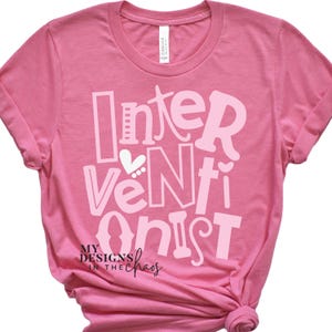 May include: Pink t-shirt with white text that reads "Interventionist" with a heart and dots above the "N". The text "My Designs in the Chaos" is below the main text.