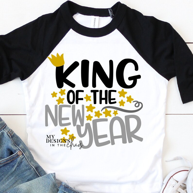 May include: White and black raglan t-shirt with the text "King of the New Year" in black and gray lettering with gold stars and a gold crown. The text "My Designs in the Chaos" is in smaller lettering below the main text.