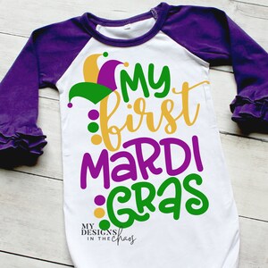 Könnte beinhalten: Weißer Langarm-Strampler mit lila und grünen Ärmeln. Der Strampler hat eine lila, grüne und goldene Narrenkappe mit dem Text "My first Mardi Gras" in Gold, Lila und Grün.