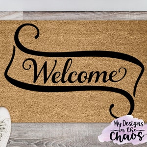 Puede incluir: Una alfombra de coco marrón con la palabra "Welcome" en una fuente de letra cursiva negra estilizada en una pancarta.