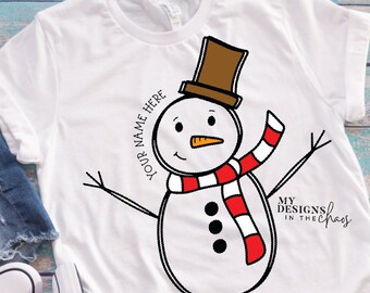 May include: White t-shirt with a cartoon snowman wearing a brown hat and a red and white scarf. The text "YOUR NAME HERE" is written around the snowman. The text "MY DESIGNS IN THE CHAOS" is written in the bottom right corner.
