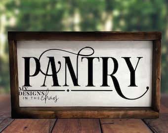Op de afbeelding: Een wit houten bord met een bruin frame, waarop het woord "PANTRY" in zwarte cursieve letters staat. Het bord bevat ook de tekst "MY DESIGNS IN THE CHAOS" in kleinere zwarte letters.