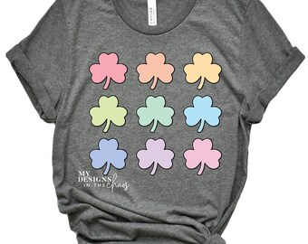 May include: A gray t-shirt with a rainbow pattern of shamrocks. The text "MY DESIGNS IN THE CHAOS" is printed on the bottom left of the shirt.