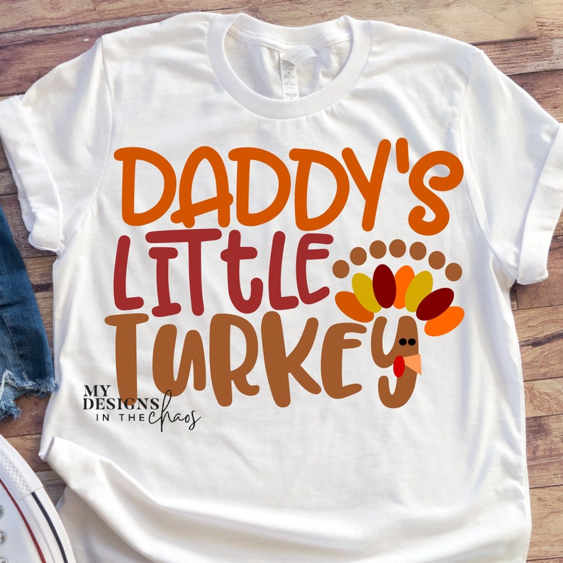 Puede incluir: Camiseta blanca con el texto "Daddy's Little Turkey" en letras naranja, rojo y marr&oacute;n. Un pavo de dibujos animados con una papada roja y plumas naranja, amarillo y rojo est&aacute; a la derecha del texto. El texto "My Designs in the Chaos" est&aacute; en la esquina inferior izquierda.