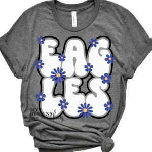 May include: Gray t-shirt with the word "EAGLES" in white, outlined in black, and decorated with blue and yellow flowers. The shirt is made of a soft material and has rolled-up sleeves.