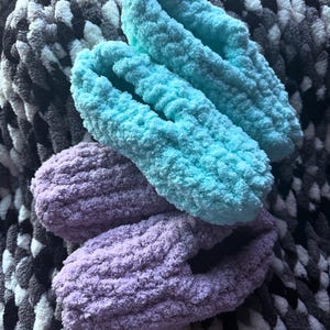 May include: Two pairs of cozy, handmade slippers. One pair is a vibrant turquoise, and the other is a soft lavender. The slippers are made of a thick, plush yarn, perfect for indoor wear. The background is a textured blanket.